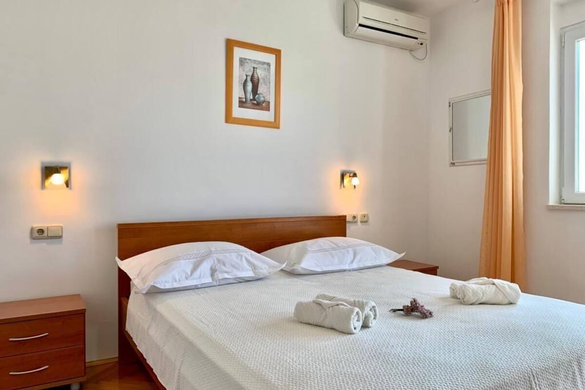 Een net opgemaakt bed met witte lakens, twee nachtkastjes, een wandairconditioner en een ingelijst schilderij boven het bed.
