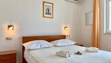 A neatly made bed with white linens, two bedside tables, a wall-mounted air conditioner, and a framed picture above the bed.