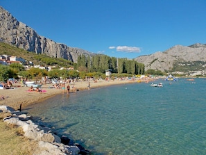 En strand med klart vatten, en stenig kust och berg i bakgrunden.