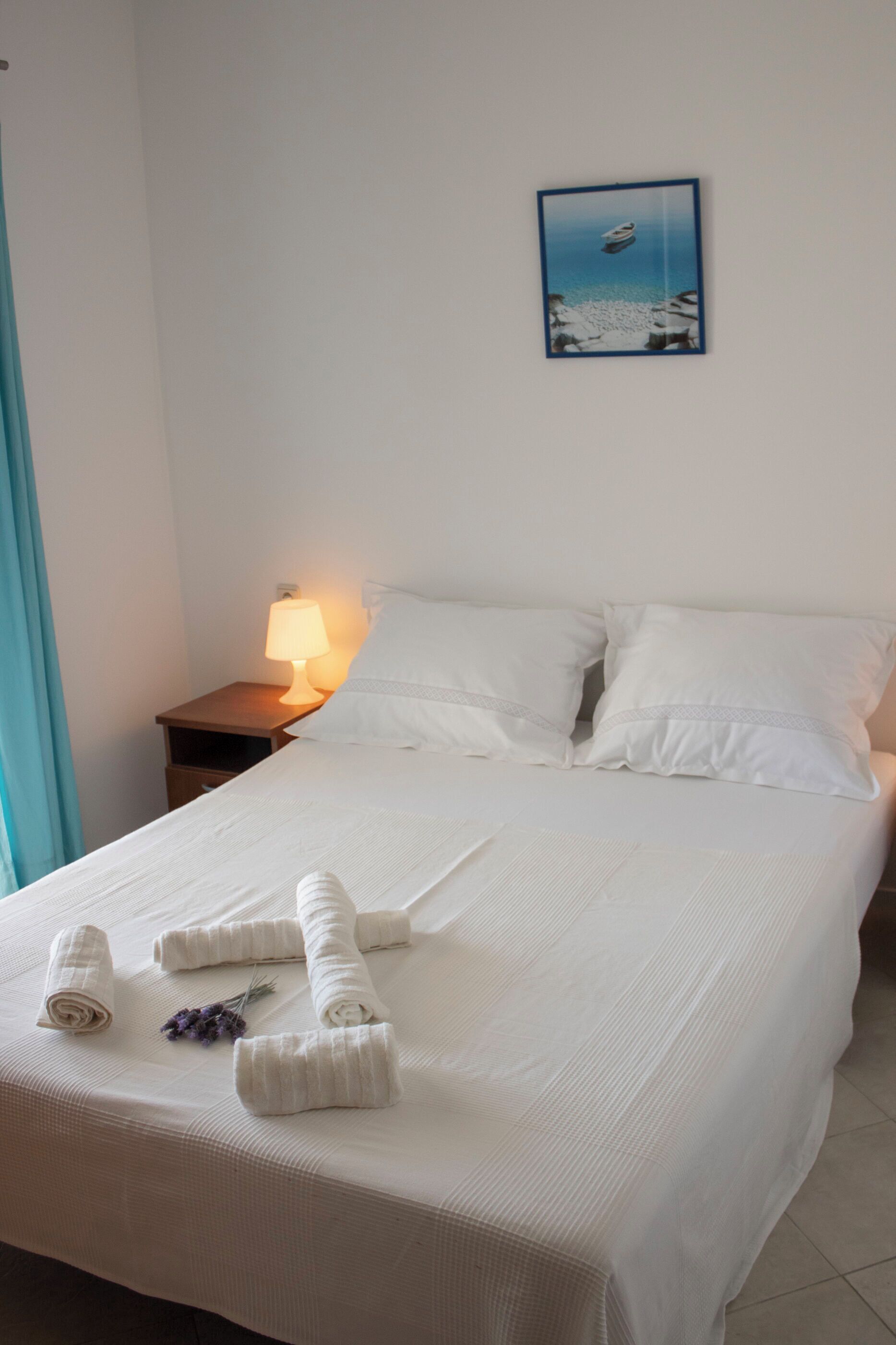 A neatly made bed with white linens and towels, a bedside lamp, and a framed picture on the wall.