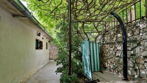 Un sentiero stretto che conduce a un muro di pietra con una struttura verde e bianca a righe, probabilmente un capanno da giardino, e un tubo di metallo a destra.
