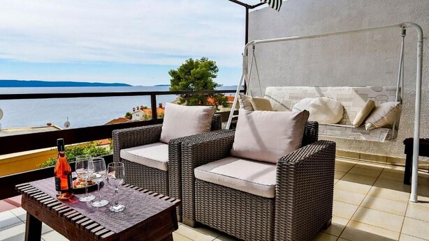 Un balcone con mobili in vimini, un tavolo con bicchieri e una bottiglia, e vista sul mare.