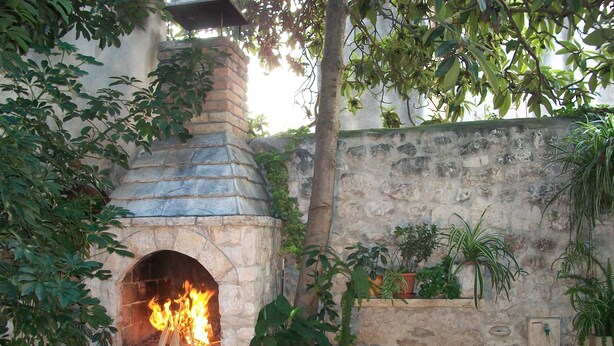 A stone-built outdoor fireplace with a fire burning inside, surrounded by potted plants and a rustic stone wall. - Apartment and Room Ivica (Trogir)