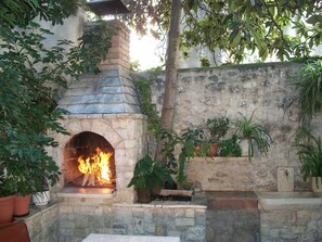 A stone-built outdoor fireplace with a fire burning inside, surrounded by potted plants and a rustic stone wall. - Apartment and Room Ivica (Trogir)