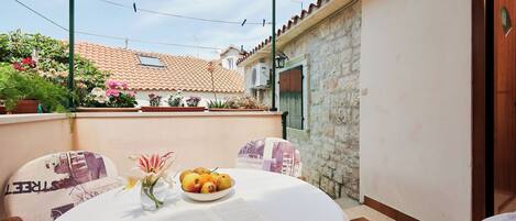 A round table with a book and a bowl of fruit, a chair with a patterned cushion, and a view of a building with a tiled roof.