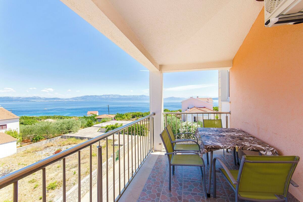 A balcony with a table and chairs, overlooking a coastal landscape with mountains and a body of water.