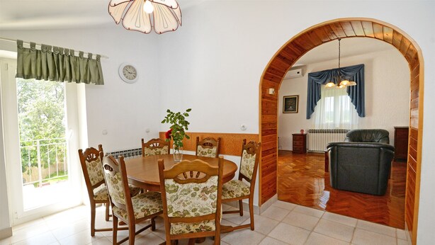 Zona pranzo con tavolo e sedie in legno, una pianta in vaso e vista attraverso un arco su un'altra stanza con termosifone e poltrona.