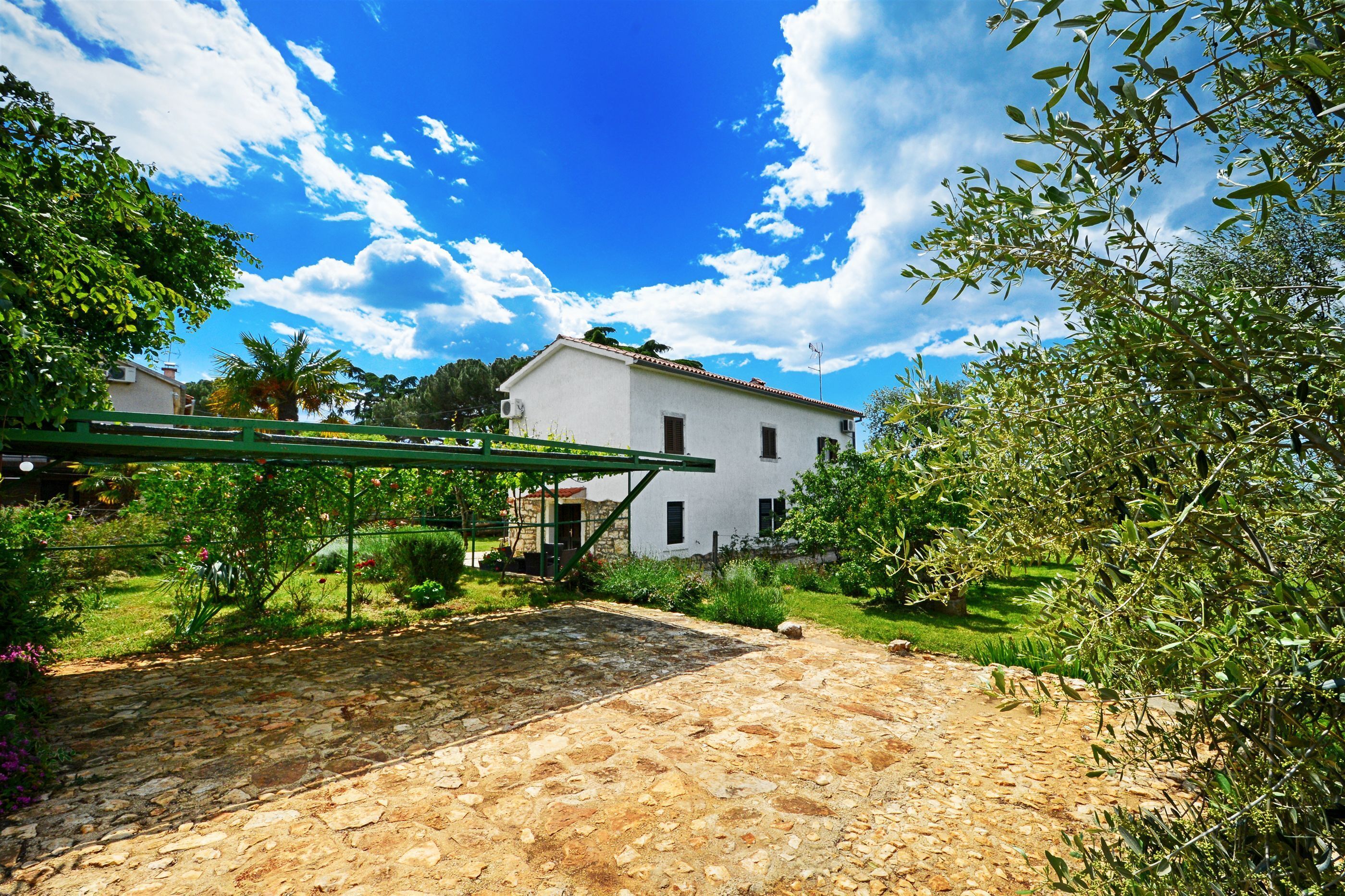A stone patio with a pergola, a white building in the background, and lush greenery.