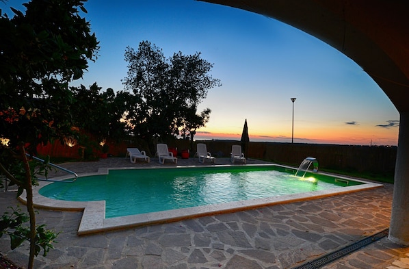 A swimming pool with a stone deck, surrounded by lounge chairs and trees, under a clear sky.