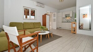 A modern living room with a green sofa, a wooden chair, and a small round table. There is a kitchen area with white cabinets and a dining table with chairs.