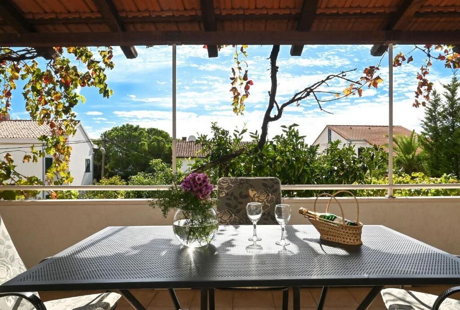 An outdoor patio with a table set for two, featuring a vase with flowers, two wine glasses, and a basket.