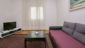 A living room with a flat-screen TV on a white stand, a black coffee table, a purple sofa, a patterned rug, and a large window with white curtains. - Apartment Zlatko (Pirovac)