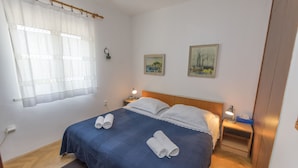 A neatly made bed with two rolled towels, a bedside lamp, and a small table with a tissue box. - Apartments Ziva (Primosten)