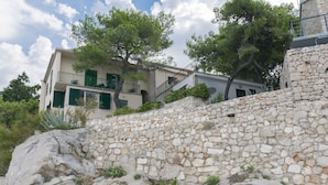 A stone wall with a building behind it, featuring a balcony with green shutters. - Apartments Ziva (Primosten)