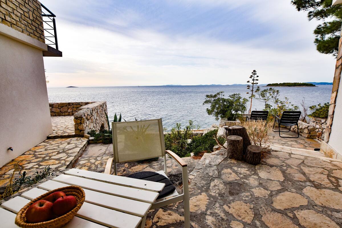 A stone patio with a white table, a wicker basket with apples, and a chair overlooking a body of water.