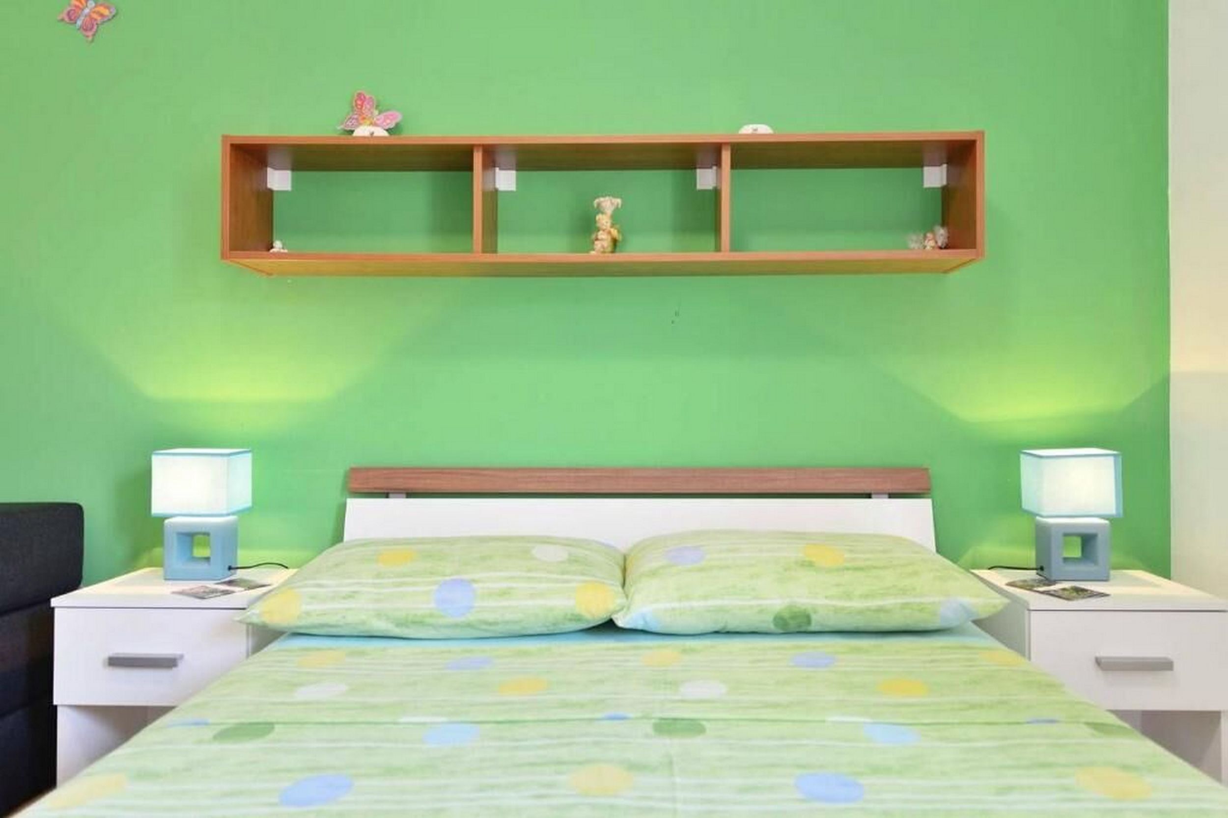 A bedroom with a green wall, a bed with a green patterned bedspread, two bedside lamps, and a wooden shelf above the bed.