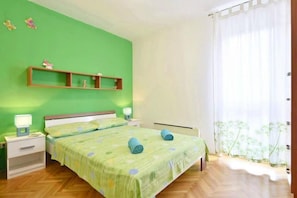 Une chambre avec un mur vert, un lit recouvert dâune couverture verte, deux tables de chevet avec des lampes, une petite table en bois et une fenĂȘtre ornĂ©e de rideaux lĂ©gers.