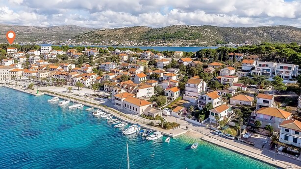 An aerial view of a coastal town with buildings, a marina, and a clear blue sea.