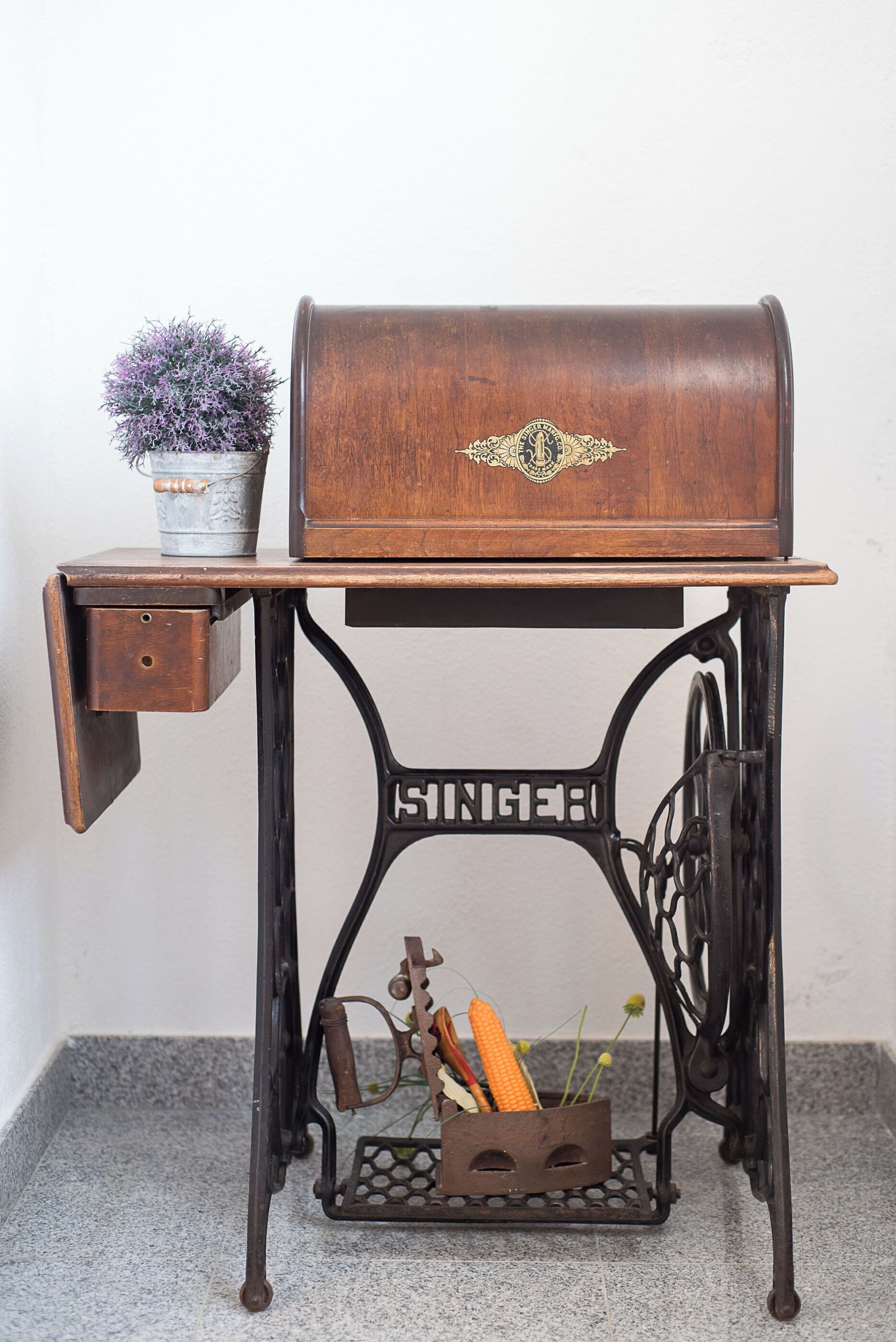 Une table ancienne pour machine à coudre, avec un plateau en bois, un tiroir et un récipient en métal contenant un épi de maïs et des fleurs.
