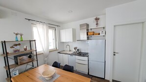 Eine kleine Küche mit weißen Schränken, einer Spüle und einem Kühlschrank. Ein Esstisch mit Stühlen und einer Schüssel steht daneben.