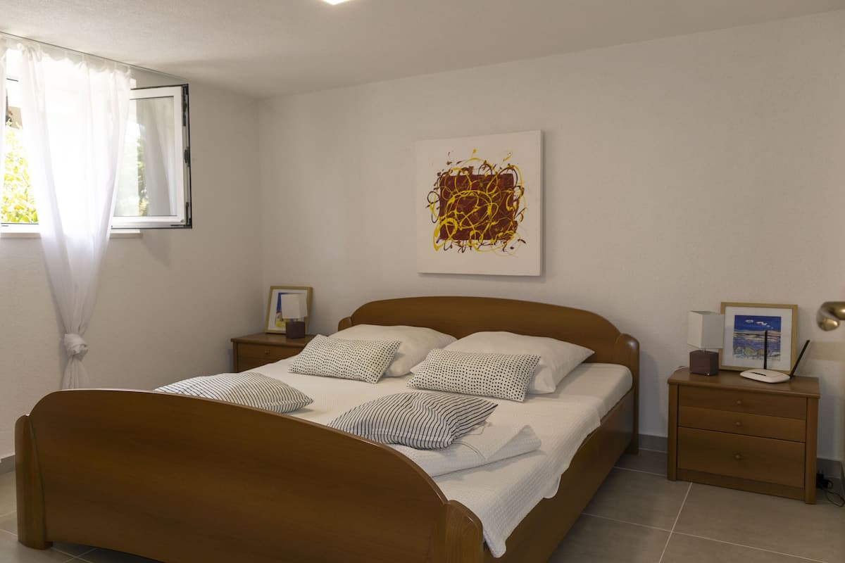 Een slaapkamer met een houten bed, wit beddengoed, een nachtkastje met een lamp en een lijst met een afbeelding aan de muur.