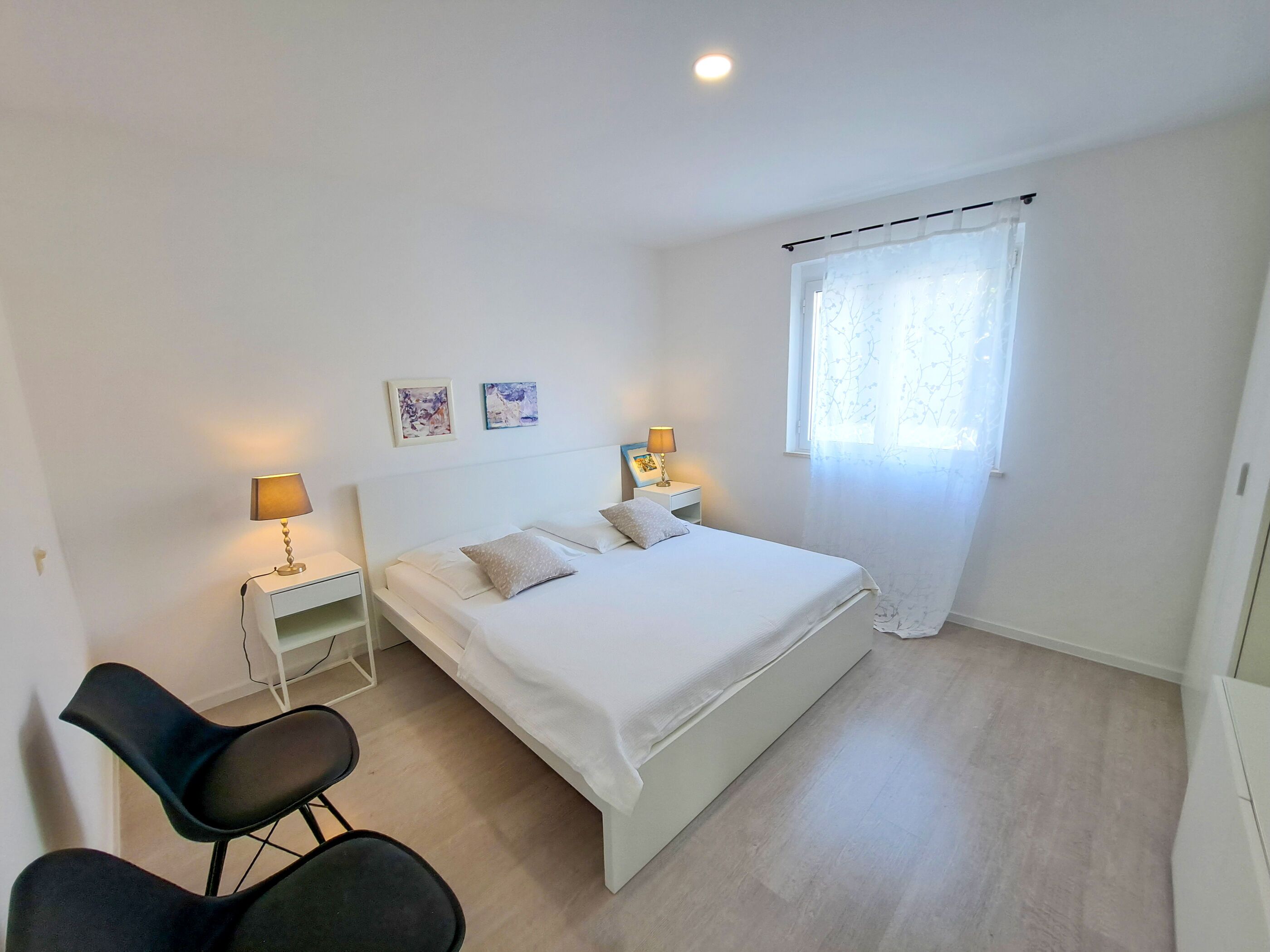 A minimalist bedroom with a single bed, bedside tables, a chair, and a window with sheer curtains.