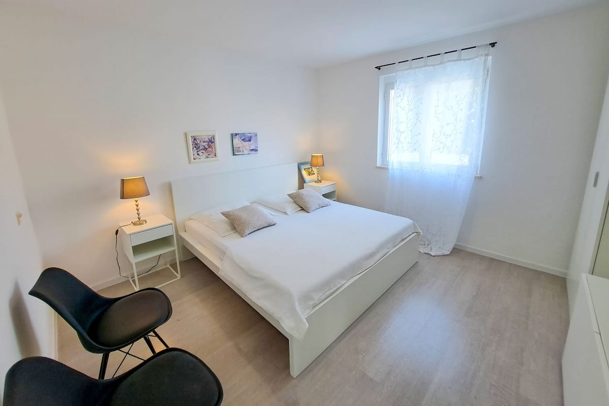 Een minimalistische slaapkamer met een enkel bed, nachtkastjes, een stoel en een raam met transparante gordijnen.