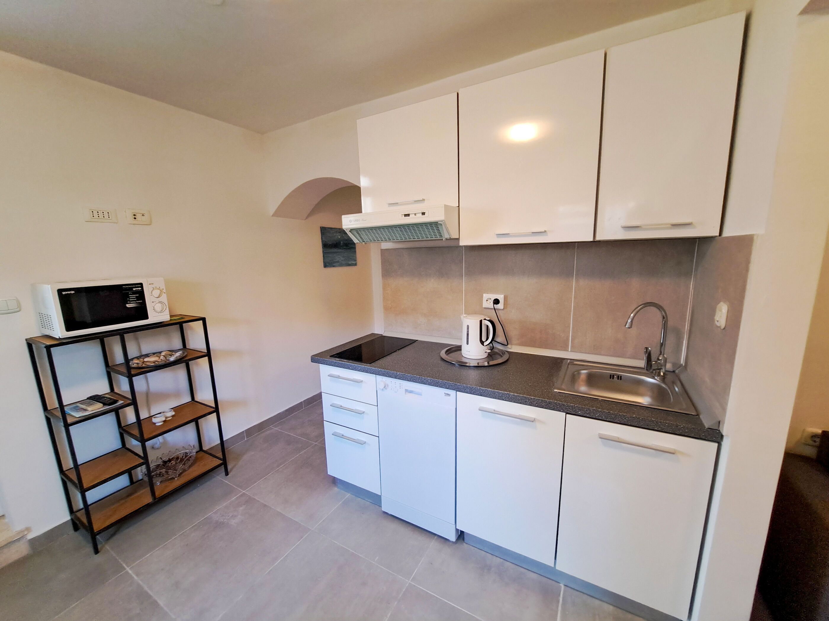 A modern kitchen with white cabinets, a black countertop, a built-in microwave, a kettle, and a stainless steel sink.