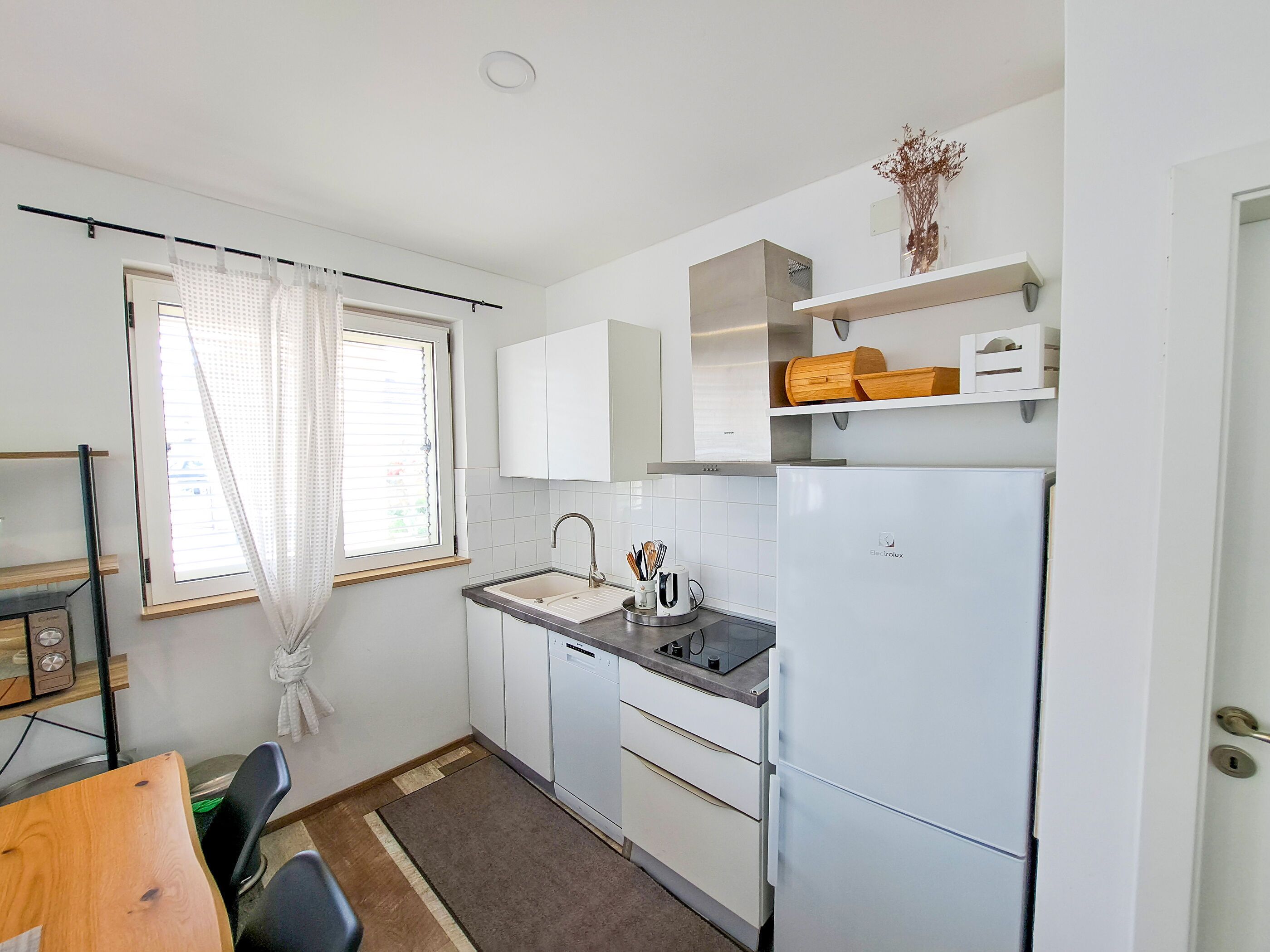 A compact kitchen with a built-in dishwasher, a sink, and a refrigerator. There is a window with curtains, a small shelf with decorative items, and a dining area with a table and chairs.
