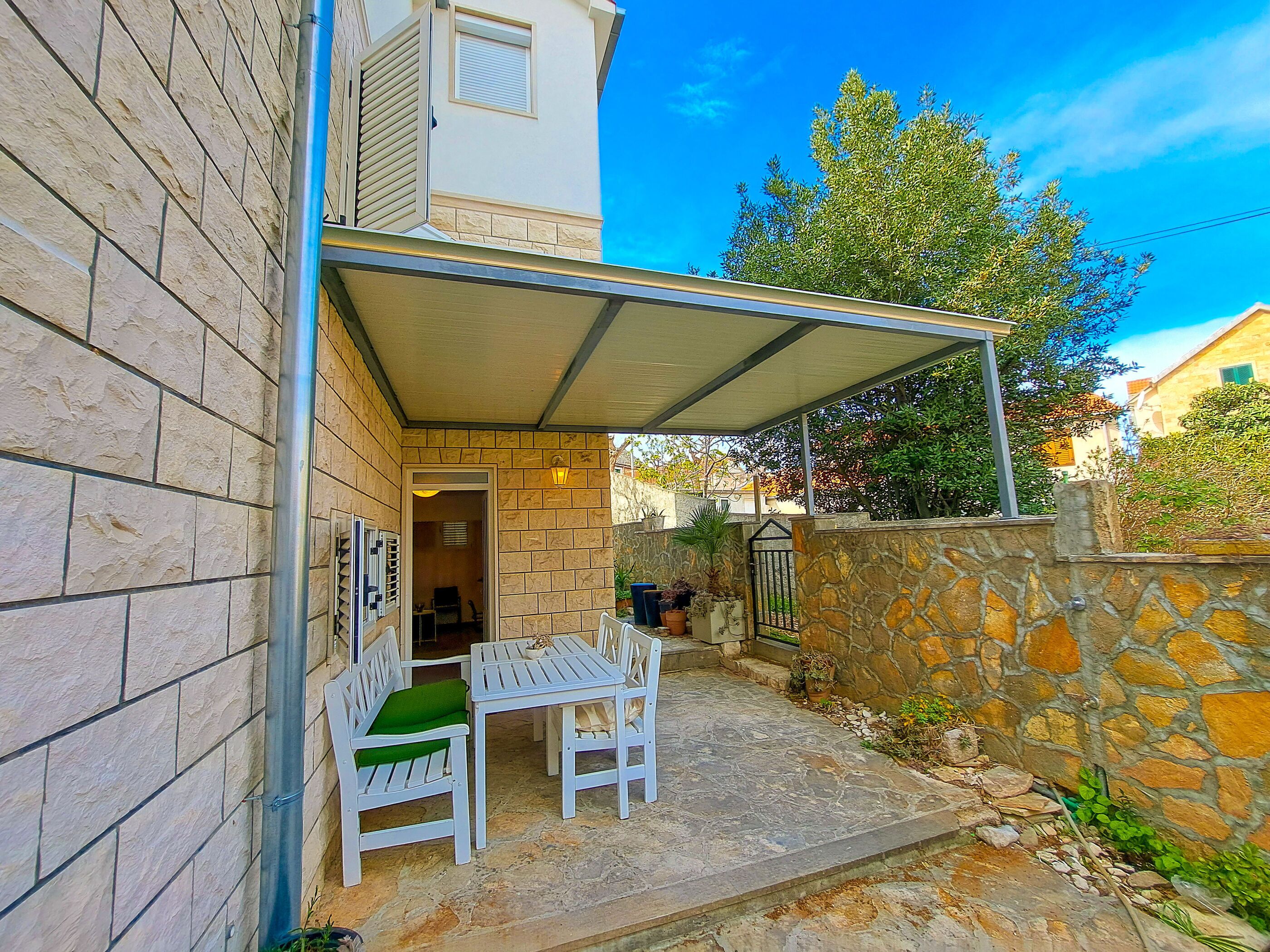 A covered outdoor seating area with stone walls and a metal roof, adjacent to a building with a door and window.