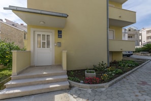 A yellow building with a white door and steps leading to a small garden with flowers. - Apartment Dora (Duce)
