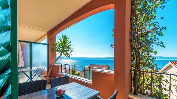 A balcony with a table and chairs, a potted plant, and a view of the sea. - Apartments Kunac (Podgora)