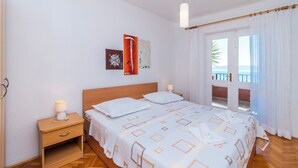 A hotel room with a bed, bedside tables, a nightstand, a window with curtains, and artwork on the wall. - Apartments Kunac (Podgora)