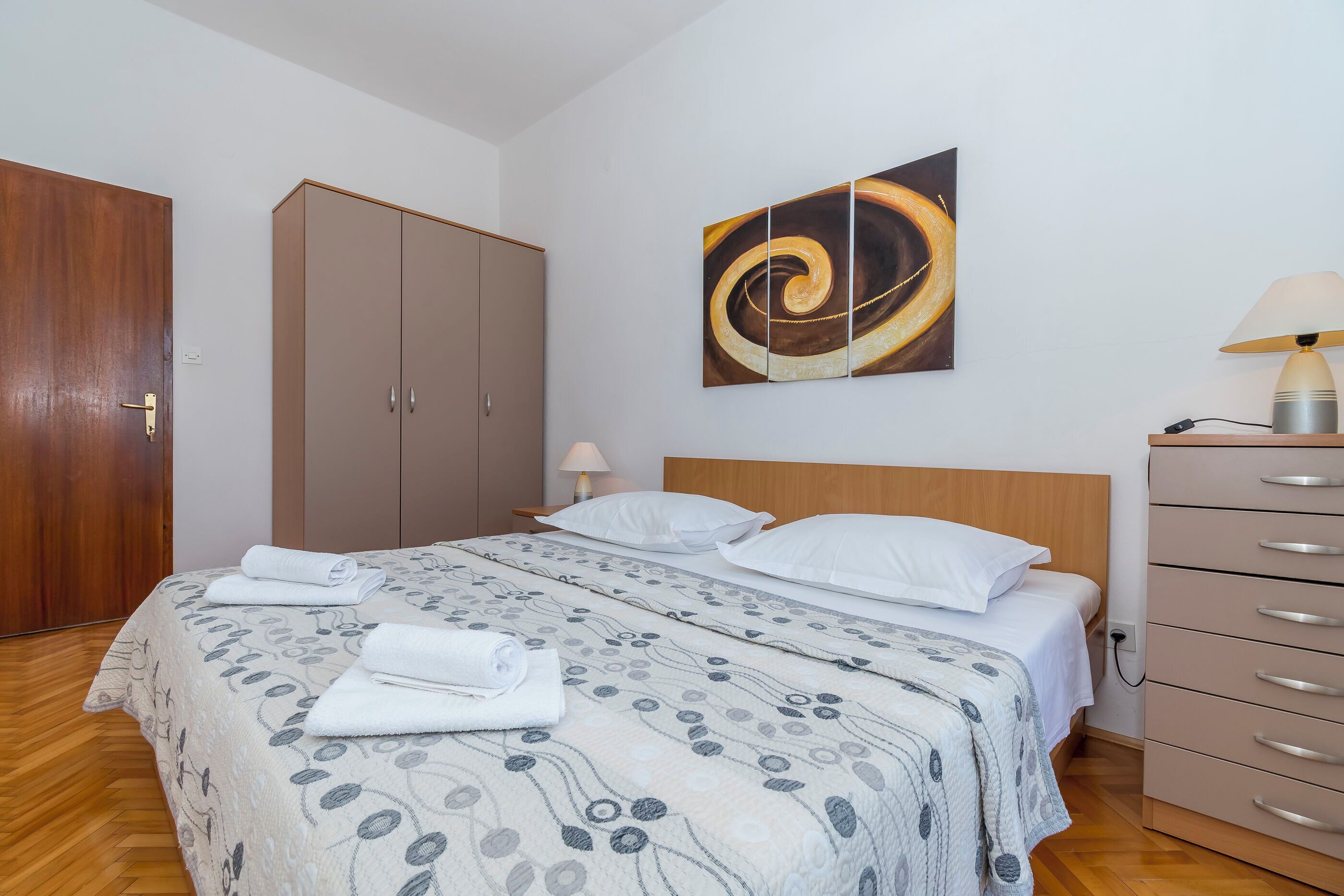 A neatly made bed with a patterned bedspread, a wooden wardrobe, a wall-mounted abstract painting, and a bedside lamp.