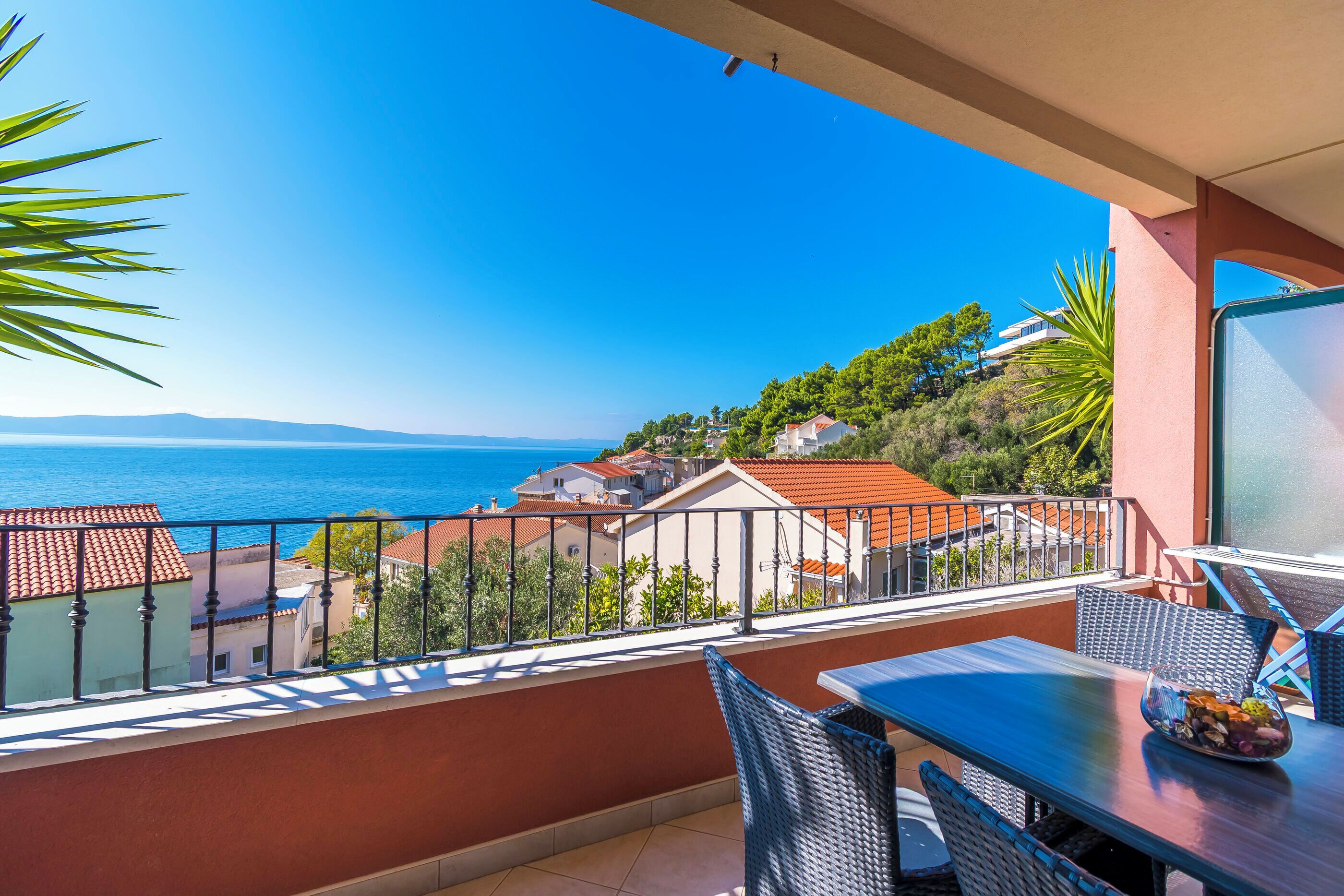 A balcony with a view of the sea, a table with chairs, and a bowl of fruit.