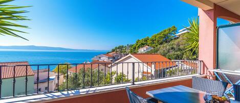 A balcony with a view of the sea, a table with chairs, and a bowl of fruit.