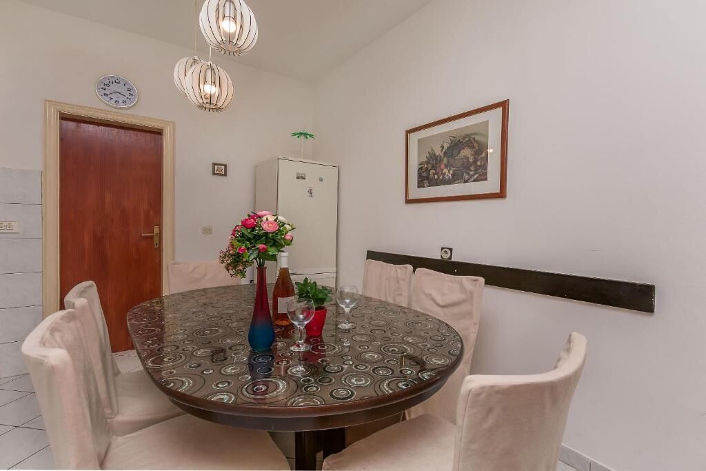 A dining area with a round table, chairs, a clock, a refrigerator, and a framed picture on the wall.