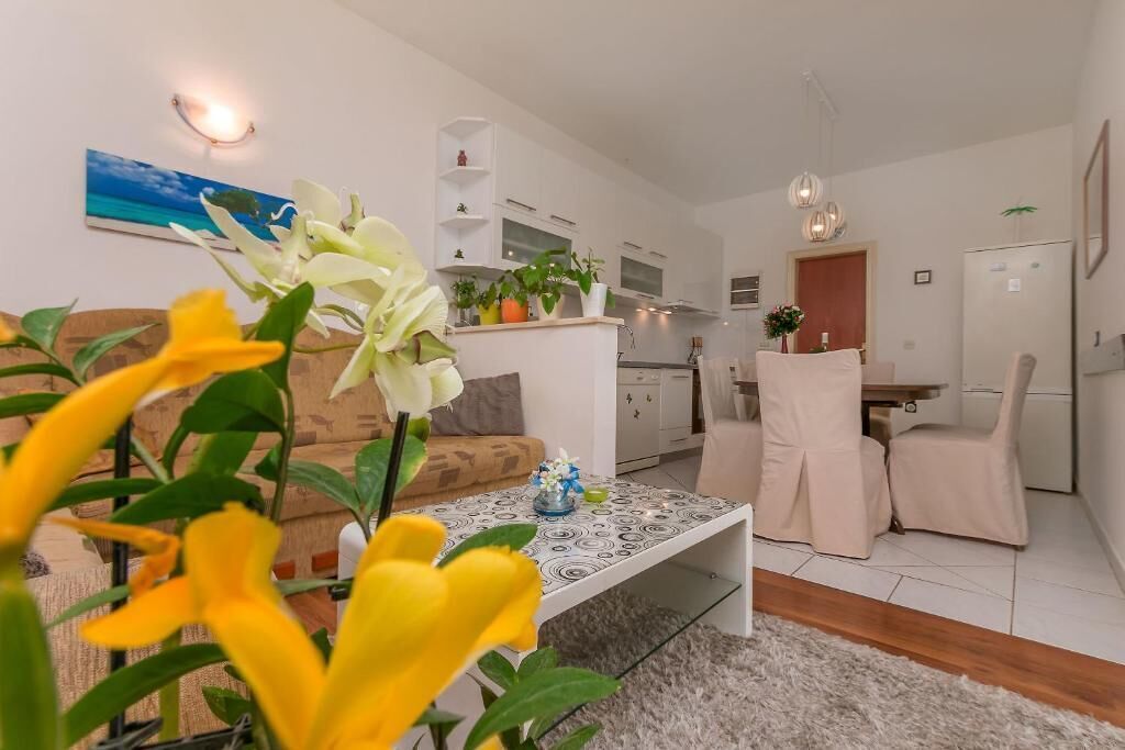 Un salon moderne avec un canapé, une table basse et un coin repas avec des chaises. On aperçoit au fond une cuisine équipée de placards et d’un réfrigérateur.