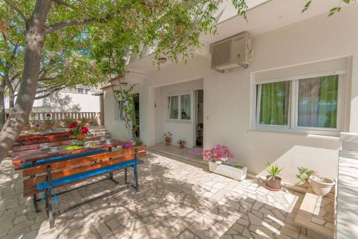 Une terrasse extérieure avec des bancs en bois, des plantes en pot et un bâtiment blanc doté de fenêtres et d’un appareil de climatisation.