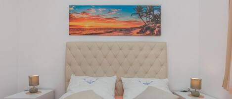 A bedroom with a large bed, two bedside tables, and a painting of a sunset over a beach.