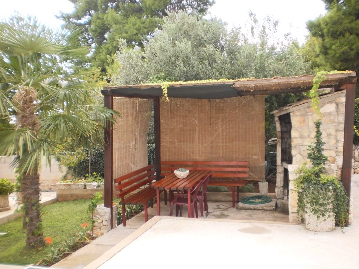 A wooden pergola with a table and benches, surrounded by greenery and a stone wall.