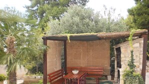 Eine Holzpergola mit Tisch und Bänken, umgeben von Grünpflanzen und einer Steinmauer.