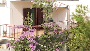 Ein weißes Gebäude mit rotem Ziegeldach, einem Balkon mit Metallgeländer und einem Baum mit violetten Blüten davor.