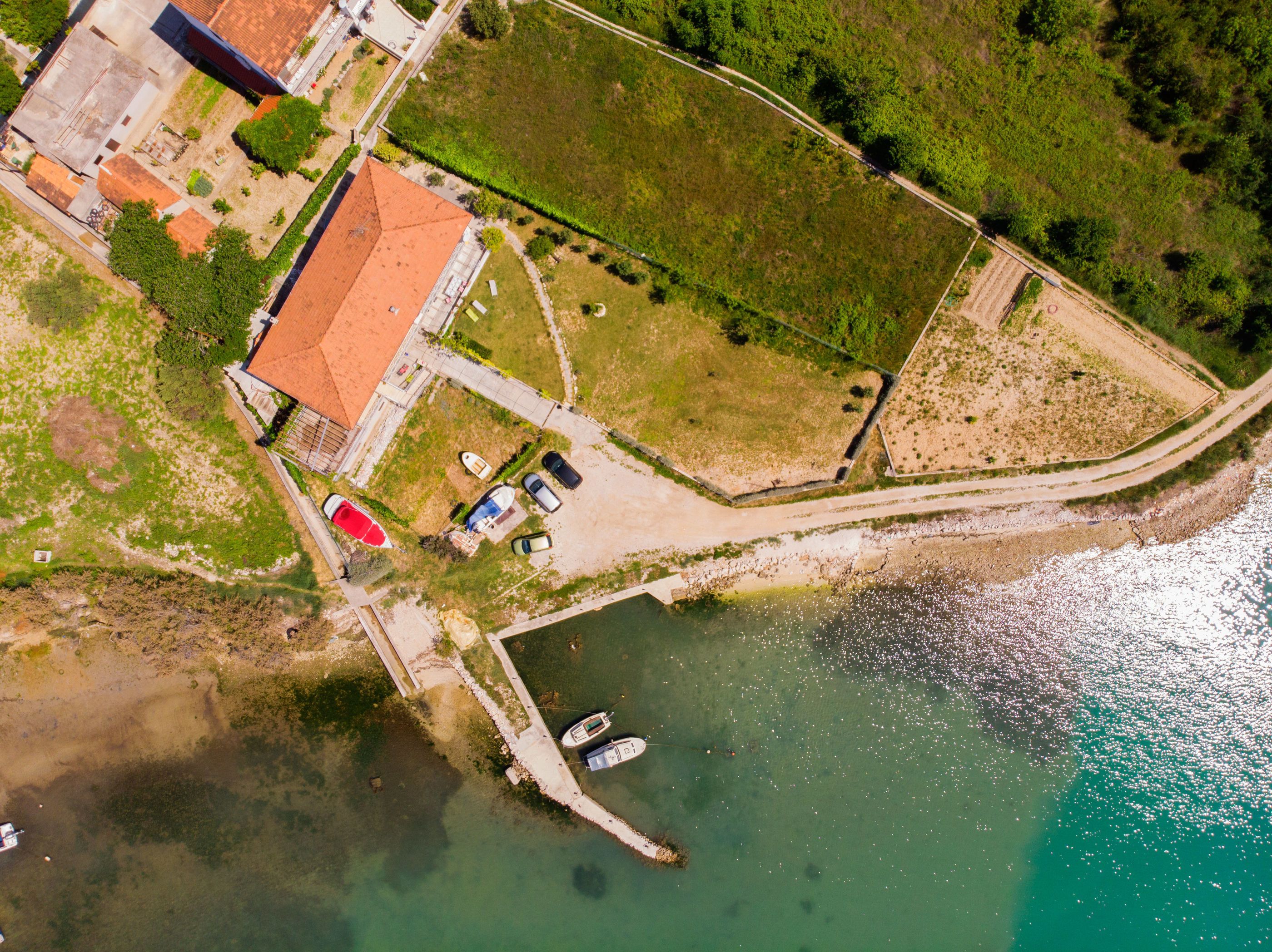Vista aérea de una propiedad costera con un edificio de techo rojo, un muelle con barcos y follaje circundante.
