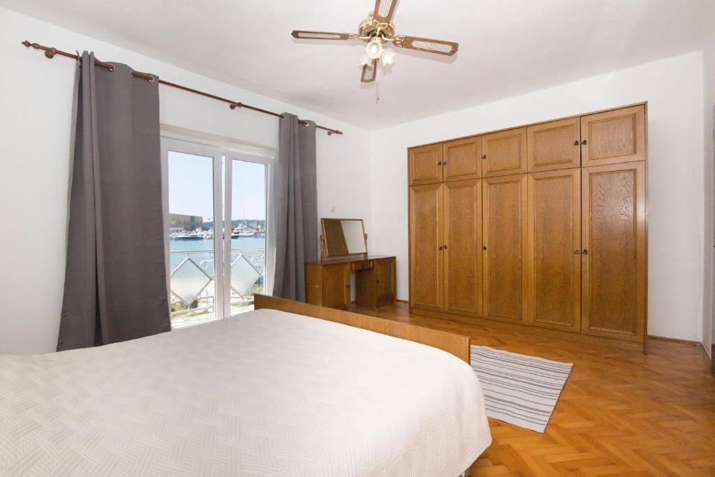 A bedroom with a bed, a wooden wardrobe, a ceiling fan, and a view of the sea through a window.