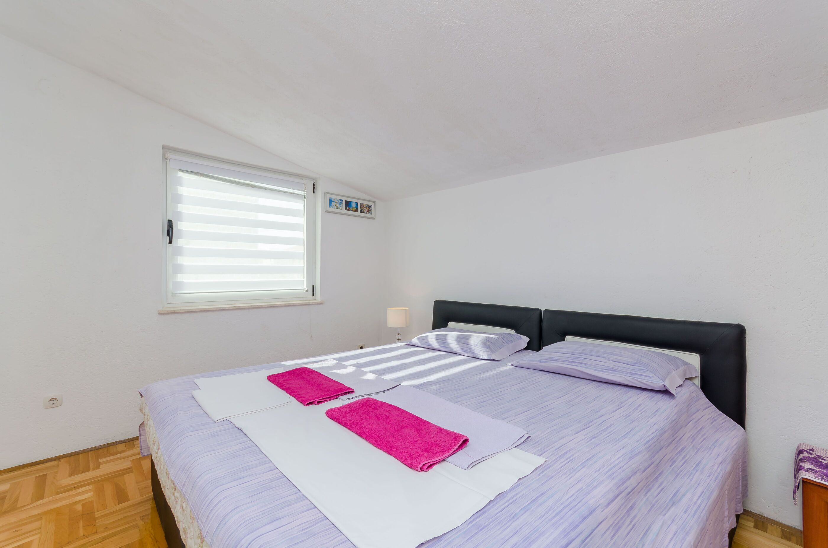 A bedroom with a bed, a window with blinds, a bedside lamp, and a small picture frame on the wall.