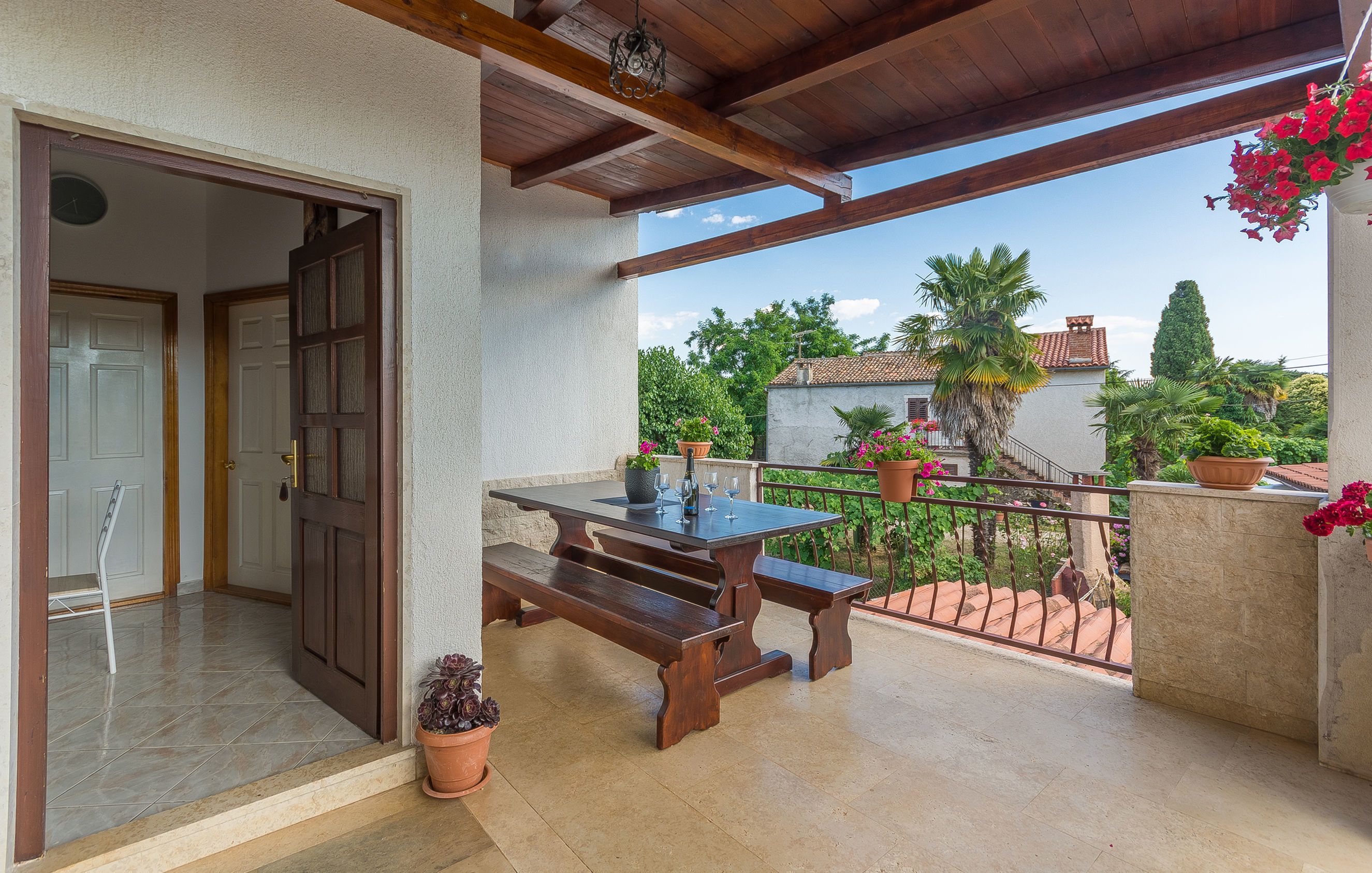 A balcony with a wooden table and benches, potted plants, and a view of a garden and buildings.