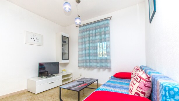 A room with a bed, a TV on a white cabinet, a small table, and a window with a curtain. - Apartment Vojka (Vrbanj)