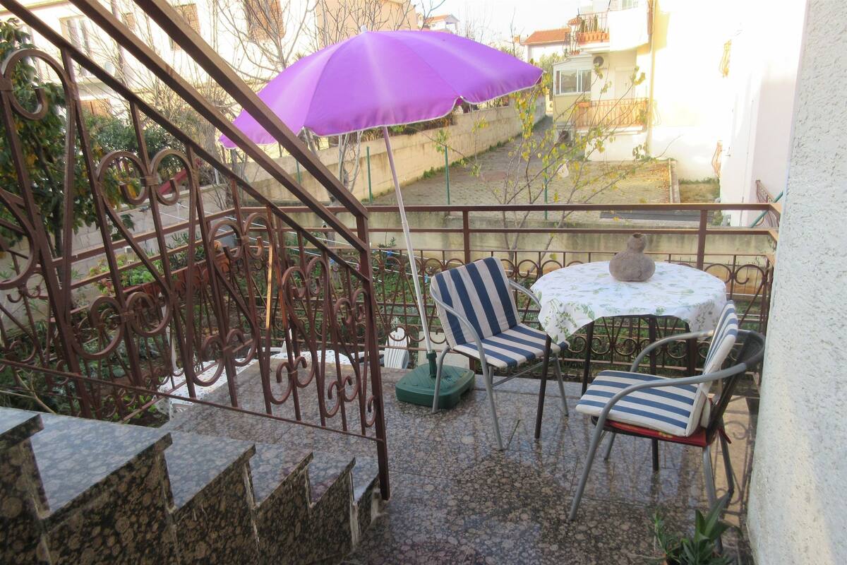 Un balcon avec un parasol violet, deux chaises à rayures, une petite table et un pot de fleurs.