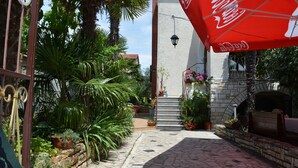 Une cour avec un chemin en pierre, des plantes en pot, un parasol à l’effigie de Coca-Cola et un mur en pierre.